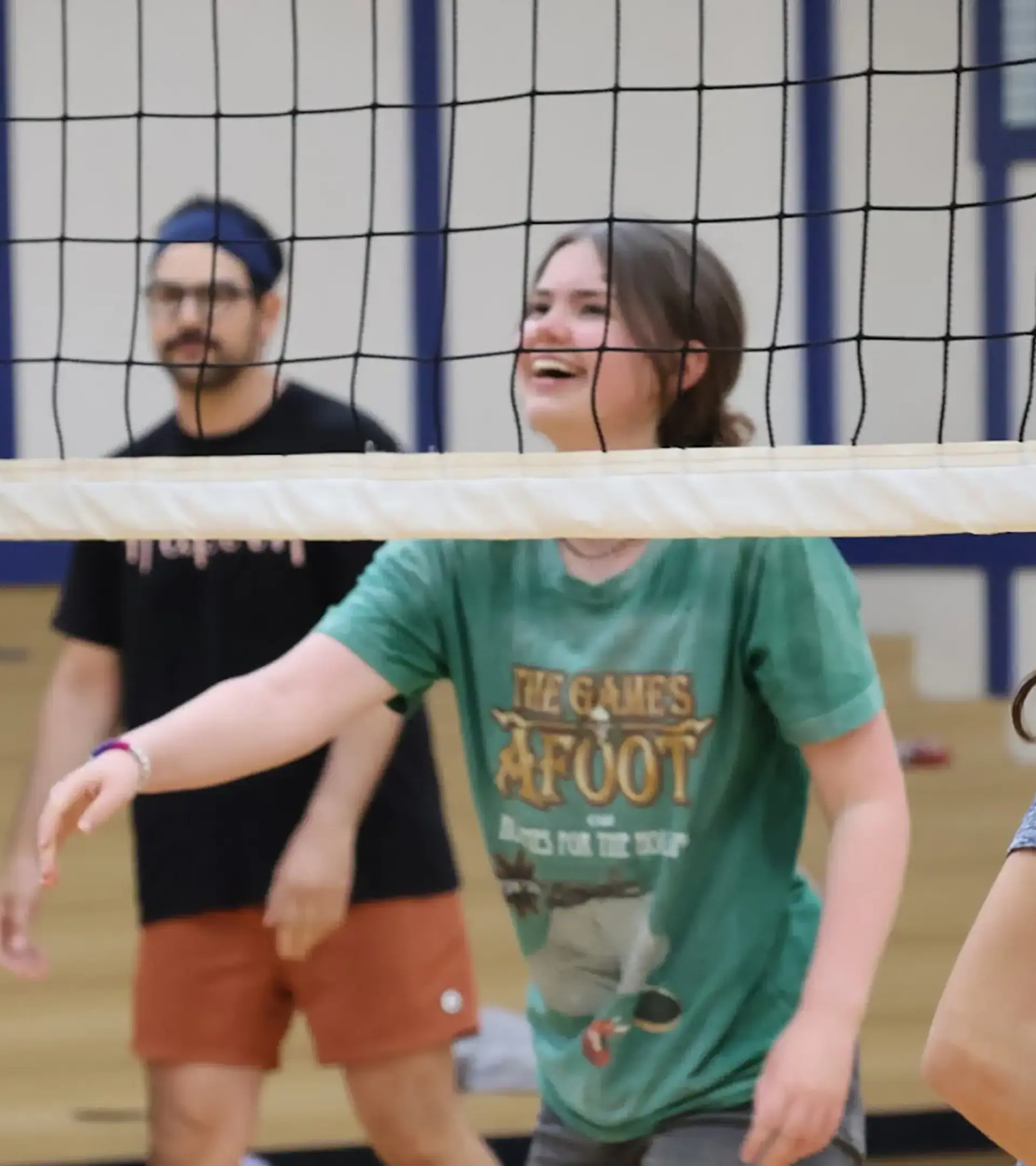 A student wearing a navy t-shirt with an Aristotle quote on the back high-fiving an adult male instructor in a blue headband, glasses, black t-shirt, and reddish shorts on a indoor gymnasium