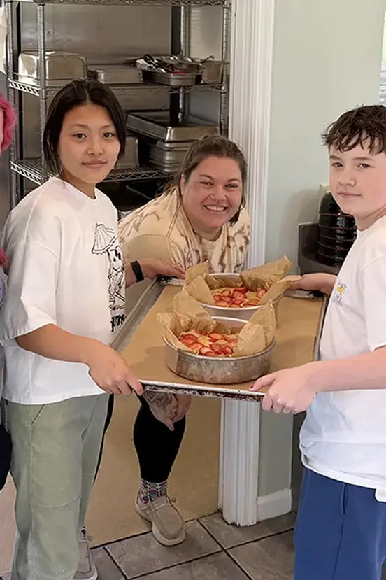 Two students and an adult carrying a tray of freshly baked pastries or food in a kitchen.