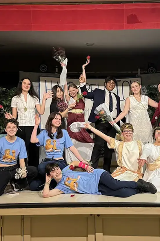 A group of students in costumes posing enthusiastically on a stage after a theater performance.