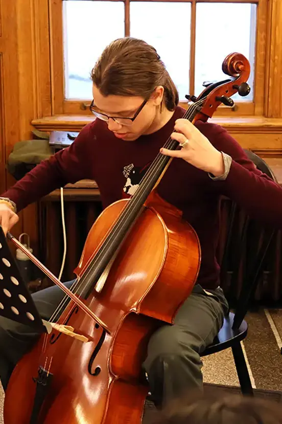 A student sitting with a cello, focused on playing music during a practice or performance. 