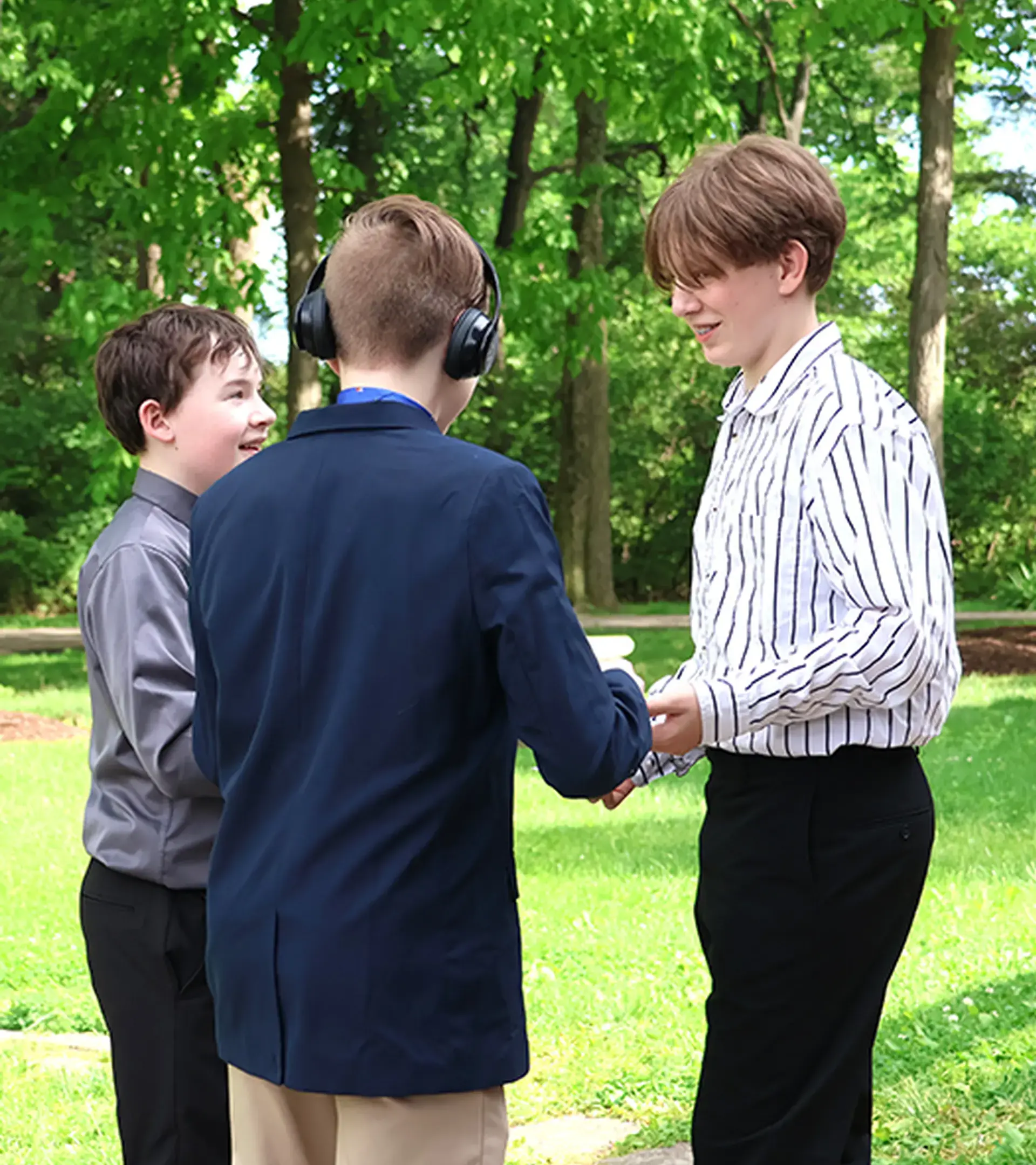 Three young male students in formal clothing talking together outdoors on a grassy lawn surrounded by trees.