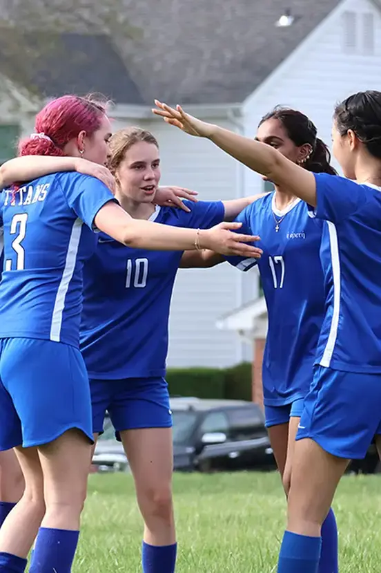Female soccer players in matching blue uniforms celebrating together on a field after scoring a goal.