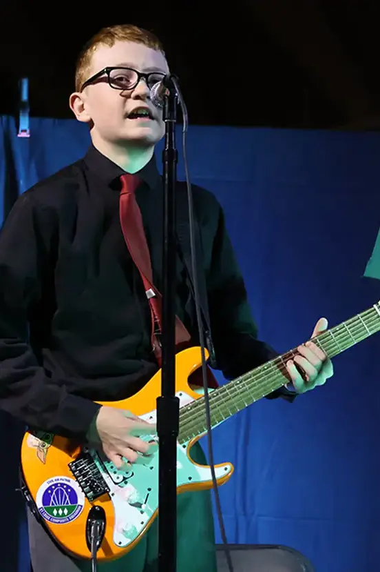 A student playing an electric guitar and singing into a microphone on stage.
