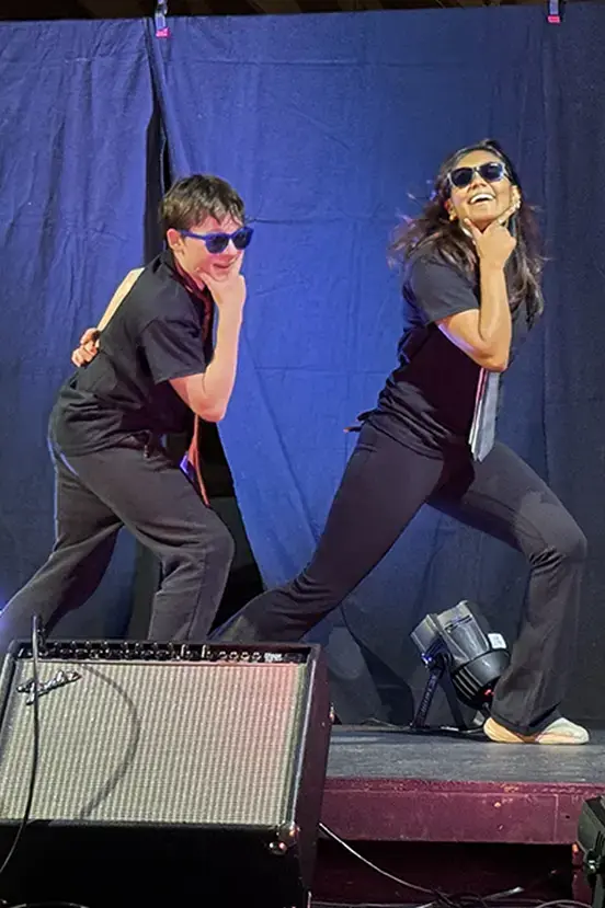 Two students in matching outfits and sunglasses posing dramatically on stage during a performance.