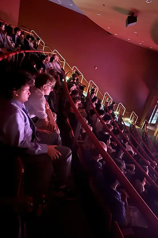 An audience of students sitting in red seats inside a dimly lit auditorium or theater.