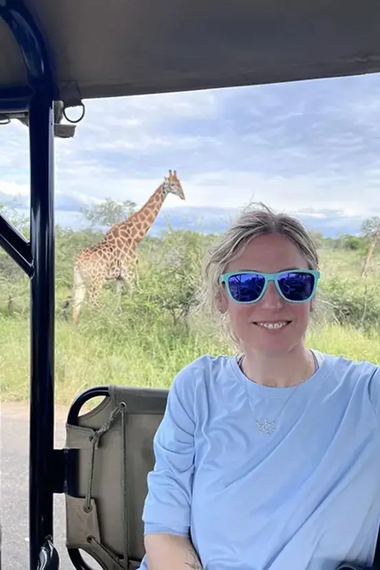 A person wearing sunglasses smiling in a vehicle with a giraffe visible in the background field. 