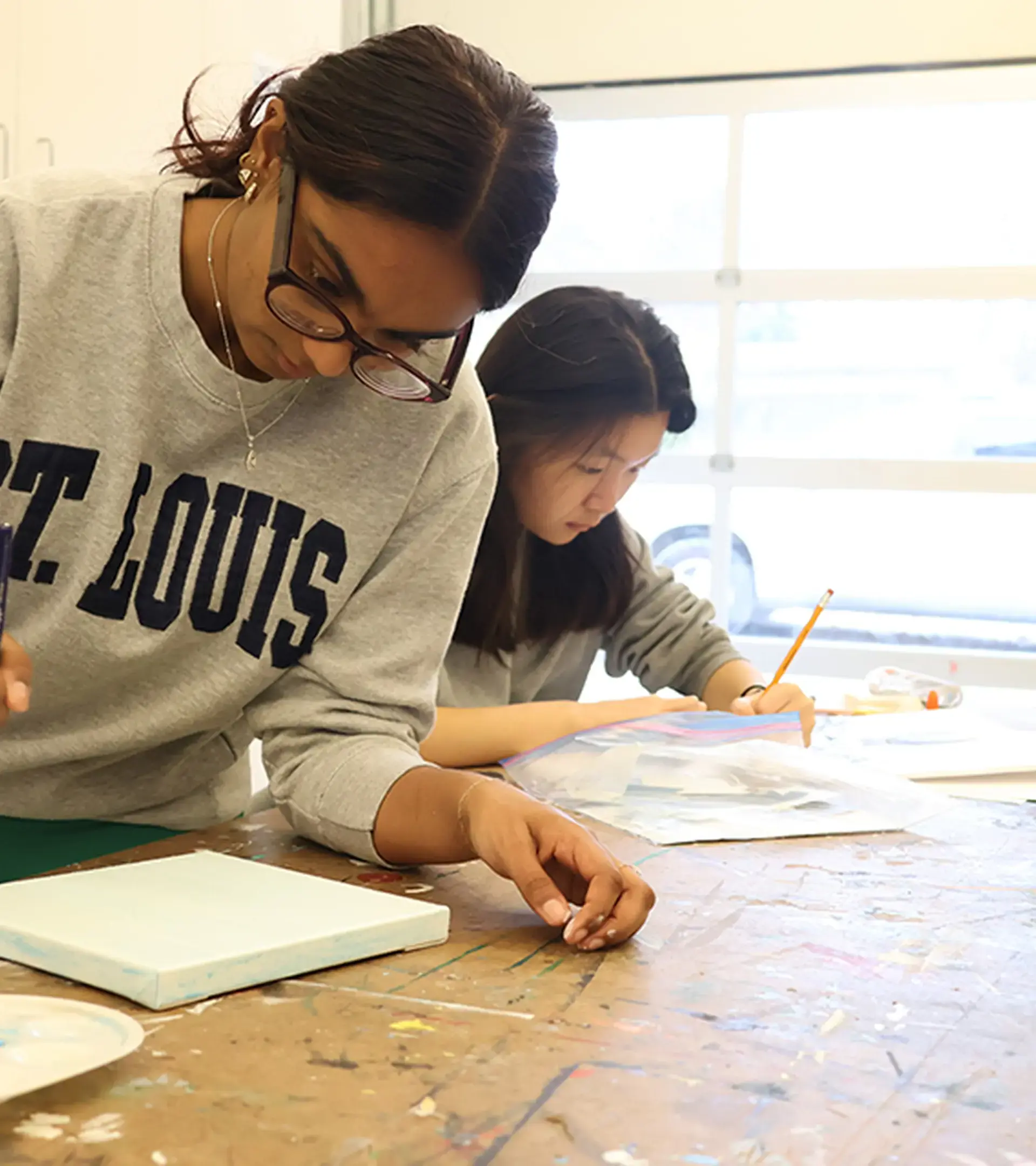 Two female students working intently on art projects at a workshop table in a bright studio.