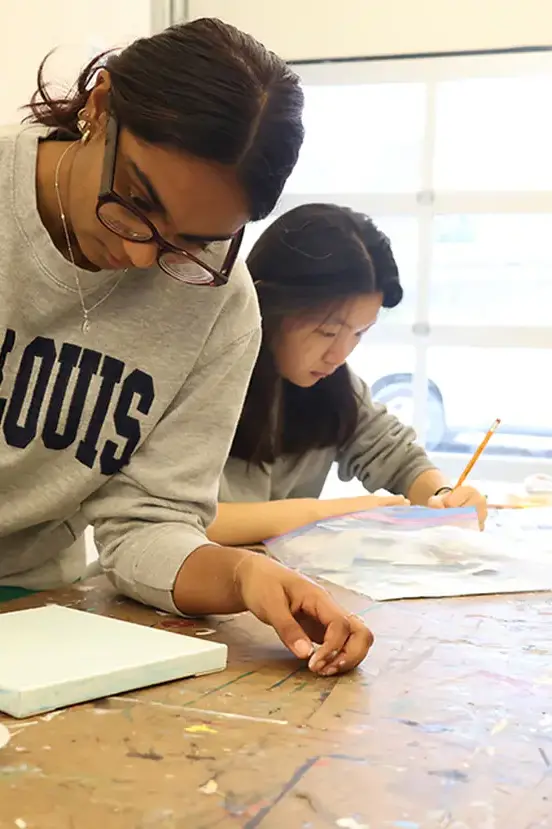 Two students working intently on artwork at a desk in an art studio or classroom.