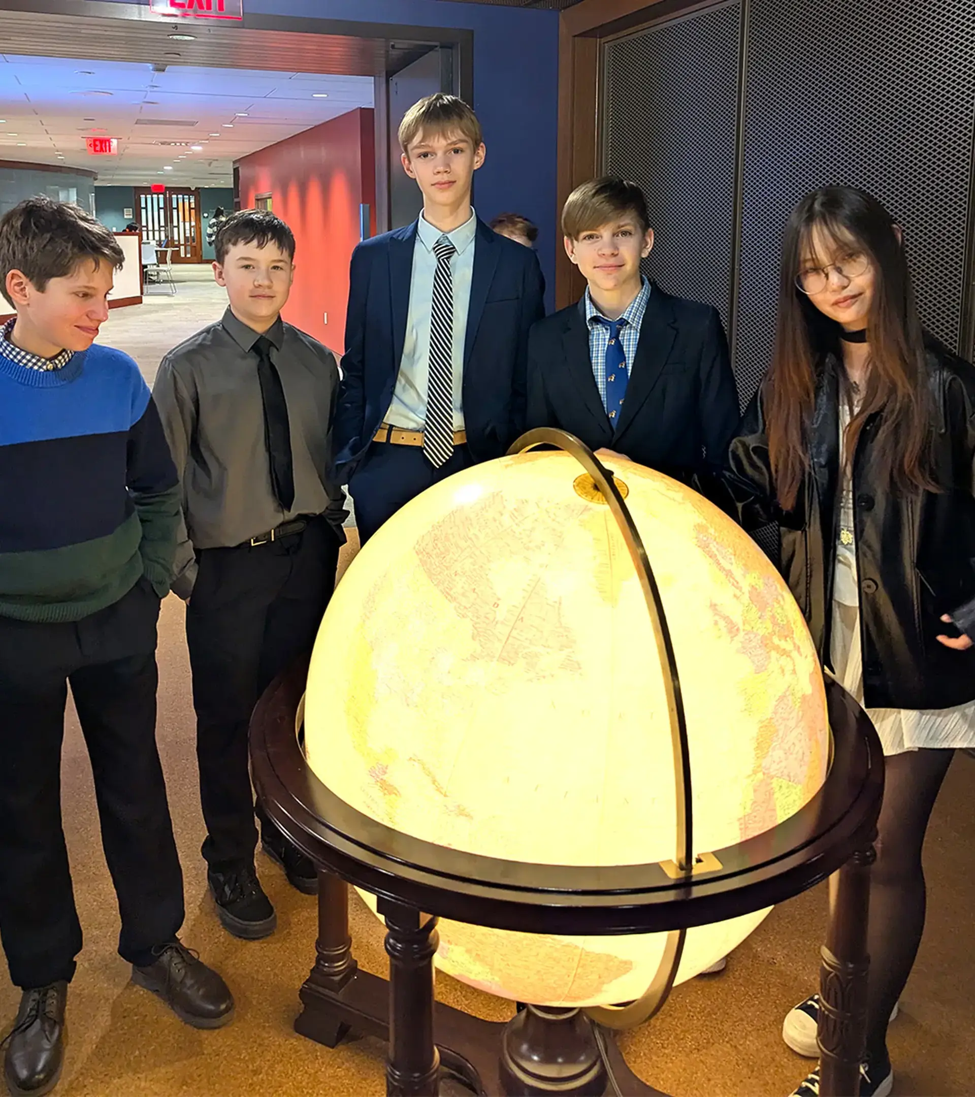 Five students in formal attire standing around a large, glowing illuminated floor globe in a hallway.